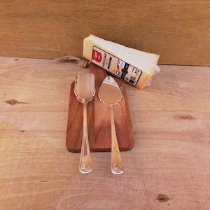 May include: A wooden cutting board with two silver cheese utensils and a wedge of Parmesan cheese. The utensils have ornate handles and are placed on the board. The cheese is wrapped in clear plastic.