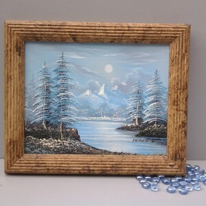 Puede incluir: Una pintura enmarcada que representa una escena de invierno nevada con &aacute;rboles azules y un lago. La pintura est&aacute; firmada "Frank E."