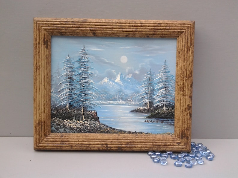 Puede incluir: Una pintura enmarcada que representa una escena de invierno nevada con &aacute;rboles azules y un lago. La pintura est&aacute; firmada "Frank E."