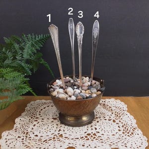 May include: Four silver spoons are arranged upright in a small, copper-colored planter filled with small, gray and white stones. The planter has a woven pattern and sits on a white crocheted doily.
