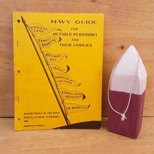 May include: A yellow booklet titled "Navy Guide for Retired Personnel and Their Families" with a black illustration of a flagpole with flags listing benefits such as medical care, family benefits, retired pay, travel, wearing of uniform, federal-state assistance, and VA benefits. A small wooden buoy painted white and burgundy is next to the booklet.