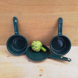 May include: Three dark green speckled enamelware pans with handles, one small frying pan, and a green and yellow artichoke on a light wood surface. The pans are empty and have a classic design.