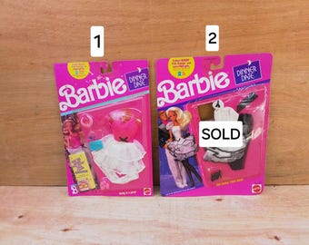May include: Two Barbie Dinner Date fashion sets in original pink packaging. Set 1 features a pink bodice with a white layered skirt and accessories. Set 2 displays a white dress with black trim and accessories, marked with a white SOLD label.