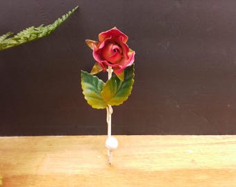 May include: A metal wall hook featuring a sculpted rose blossom in shades of red and pink, accompanied by two green leaves. The stem of the rose extends downward to form a white hook, which is finished with a small, round, white ball at the tip.