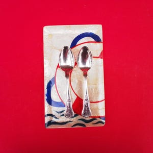May include: Two silver spoons rest on a decorative rectangular dish with a nautical design. The dish features a white background with blue and red accents, including a life preserver motif and wavy lines. The background is a solid red.
