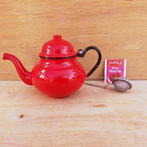 May include: A small, red teapot with a black handle and lid, next to a tea infuser and a tea bag. The teapot is on a wooden surface. The tea bag reads "IMPRA Rose Black Tea."