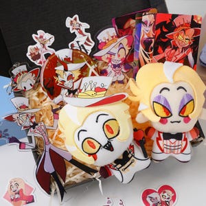 May include: A box filled with character merchandise, including plush dolls, acrylic charms, and trading cards. The plush dolls have yellow hair and red accents. The acrylic charms and cards feature various character designs in vibrant colors.