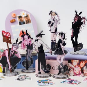 May include: Acrylic anime character figurines with bunny ears. The figures are in various poses and outfits, with a cafe sign in the background. The color palette includes black, white, pink, and red.