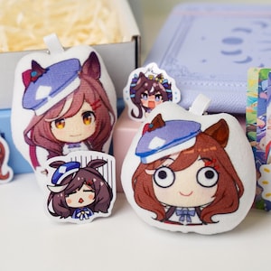 May include: A collection of anime-style character plush keychains and stickers. The plush keychains are white with a cartoon character wearing a blue hat and bow. The stickers feature the same character in various poses.