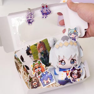 以下が含まれることがあります： 透明な窓が付いた白いギフトボックス。ぬいぐるみ、キャラクターカード、ステッカーが入っています。ぬいぐるみは白で、青と金のアクセントがあります。箱は手で開けられています。