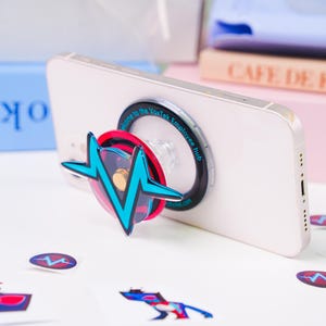 Może przedstawiać: Biały smartfon z dołączonym wysuwanym uchwytem na telefon. Uchwyt ma turkusowo-czerwony wzór w kształcie błyskawicy. Naklejki z tym samym wzorem są rozrzucone wokół telefonu.