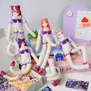 May include: A collection of anime-style plush dolls with long limbs and detailed facial features. The dolls wear white and purple sailor-style outfits. Some dolls are posed, while others are in a box with stickers and cards.