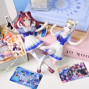 May include: Two anime-style plush dolls in sailor outfits, one with red hair and the other with white hair, are the focal point. The dolls are surrounded by stickers, cards, and a light blue notebook. The color palette includes white, blue, and pink.