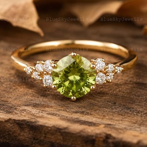 Puede incluir: Un anillo de oro con una piedra preciosa de peridoto verde lima, redonda y facetada, en el centro, flanqueada por un grupo de pequeños diamantes blancos redondos y claros a cada lado, montado en una banda de oro amarillo pulido.