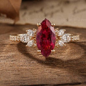 Puede incluir: Un anillo de oro con una piedra preciosa roja oscura de talla marquesa como pieza central. El anillo está adornado con piedras preciosas más pequeñas y transparentes y engastado en una banda texturizada. El anillo se exhibe sobre una superficie de madera.