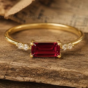 May include: A gold ring featuring a horizontal emerald-cut red gemstone at the center, flanked by two small round clear stones on each side. The band is thin and polished gold, showcasing a minimalist design with a vibrant, deep red focal point.