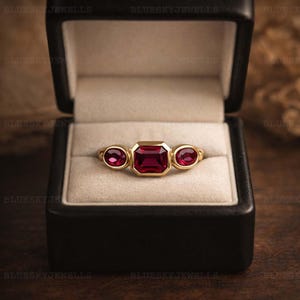 May include: A gold ring featuring a central emerald-cut ruby, with two oval rubies on either side. The ring is presented in a black box with a cream interior, highlighting the ring's design and colour.