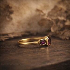 May include: A gold ring with three ruby gemstones. The ring has a rectangular central ruby, with two smaller oval rubies either side. The ring is set against a dark, textured background.