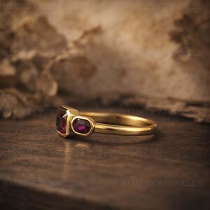 May include: A gold ring with two red gemstones. The larger stone is rectangular, the smaller is oval. The ring is displayed on a wooden surface. The gemstones are a deep red colour.