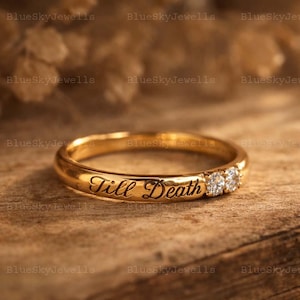 Op de afbeelding: Een gouden ring met de inscriptie "Till Death" en drie kleine diamanten. De ring ligt op een houten oppervlak, met de woorden "BlueSkyJewells" op de achtergrond.