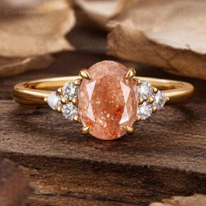 May include: A gold ring with an oval, peach-coloured gemstone at its centre. The ring is embellished with small, round, clear gemstones on either side of the main stone. The ring is set on a wooden surface.