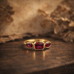 May include: A gold ring featuring three gemstones. The central stone is an emerald-cut ruby, with two oval-cut rubies either side. The ring is set on a dark wooden surface.