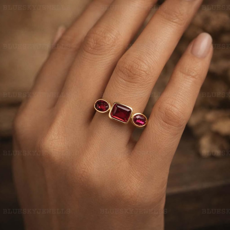 May include: A gold ring featuring three red gemstones. The central stone is rectangular, with two oval stones on either side. The ring's design is showcased against a warm, neutral backdrop, emphasizing the jewellery's colour and style.