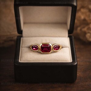 May include: A gold ring featuring a central emerald-cut ruby, with two oval-cut rubies either side. The ring is presented in a black jewellery box with a cream-coloured lining.