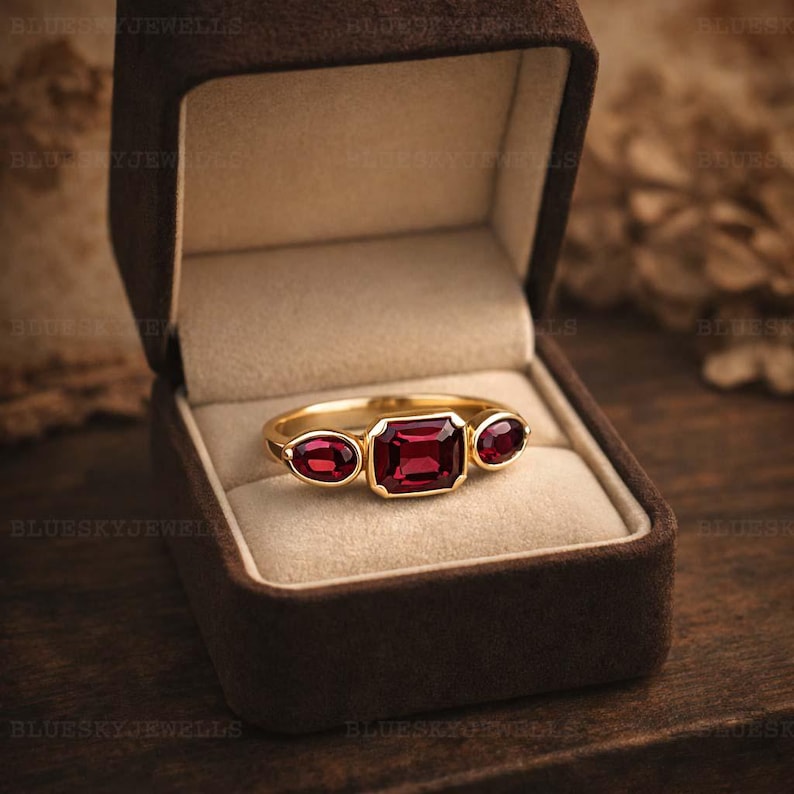 May include: A gold ring with three red gemstones, one rectangular and two oval-shaped, displayed in a brown velvet jewellery box. The ring is the focal point, with the gemstones reflecting light. This is a piece of jewellery.