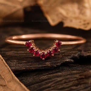 Pode incluir: Um anel de ouro rosa com uma banda em forma de V cravejada com sete pequenos rubis redondos de cor vermelha vibrante. O metal tem um acabamento liso e polido, e as pedras estão dispostas num arco simétrico no centro da banda do anel.
