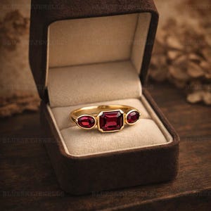 May include: A gold ring with three red gemstones, one rectangular and two oval-shaped, set in a brown velvet jewellery box. The ring is centered in the box, with the gemstones catching the light. The ring is a piece of jewellery.