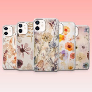 Op de afbeelding: Een verzameling van vijf transparante telefoonhoesjes met bloemmotieven. Elk hoesje heeft een ander arrangement van gedroogde bloemen in beige, bruin, oranje, paars en geel. De hoesjes staan tegen een zachtroze achtergrond.