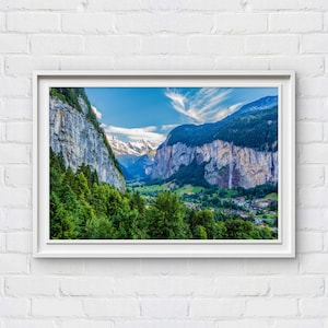 Può includere: Fotografia incorniciata di un paesaggio di valle con una cascata, montagne e un villaggio. Il cielo è blu con nuvole. Alberi verdi e vegetazione in primo piano.