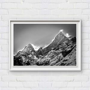 Op de afbeelding: Zwart-wit ingelijste foto van een bergketen. De toppen zijn bedekt met sneeuw en de lucht is bewolkt. De afbeelding wordt weergegeven in een witte lijst tegen een witte bakstenen muur.