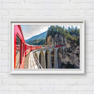 Peut inclure: Impression encadrée d'un train rouge traversant un pont en pierre. Le train suit une voie incurvée, avec une montagne et une forêt en arrière-plan. L'image est encadrée en blanc.