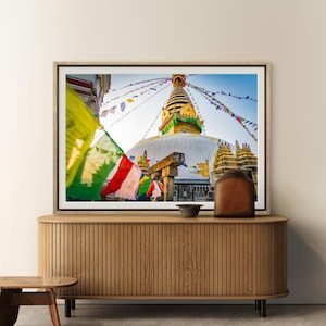 Puede incluir: Fotografía enmarcada de una estupa blanca y dorada con coloridas banderas de oración. La estupa tiene una aguja dorada y se encuentra sobre un cielo azul. La fotografía se exhibe en un mueble de madera.