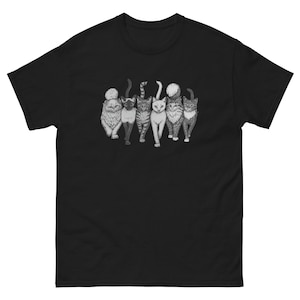 Peut inclure: T-shirt noir avec une illustration en niveaux de gris de six chats. Les chats, aux motifs de fourrure variés, sont représentés dans différentes poses. Le motif est centré sur le t-shirt, mettant en valeur les félins.