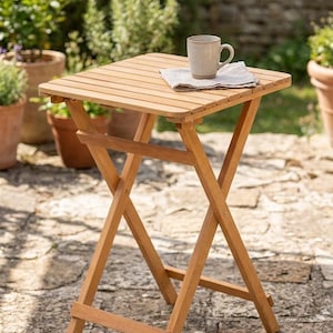 Könnte beinhalten: Kleiner, quadratischer, klappbarer Holztisch mit hellbraunem Finish. Ein beiger Becher und eine Leinen-Serviette liegen auf der Latten-Tischplatte. Der Tisch steht auf einer Steinpflasterung, im Hintergrund Grün. Ca. 40 cm x 40 cm.