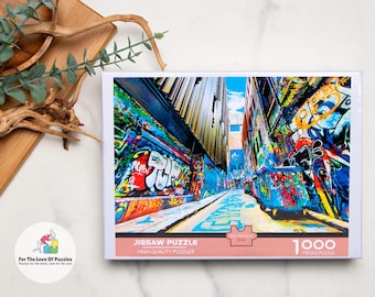May include: A 1000-piece jigsaw puzzle featuring a vibrant street scene covered in colorful graffiti art. The puzzle box displays the image of a narrow alleyway with buildings on either side, showcasing a variety of graffiti styles and colors. The text "Jigsaw Puzzle" is visible.