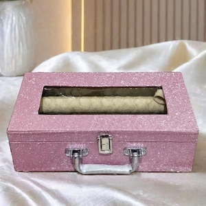 May include: A rectangular, pink glitter-covered box with a silver handle and clasp. The box has a clear window on top, revealing a rolled item inside. The box is resting on a white, silky surface.