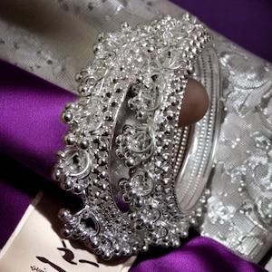 Op de afbeelding: Zilverkleurige metalen servetring met sierlijke bloemen- en kralendetails. De ring staat tegen een achtergrond van paarse en zilveren stof, met een crèmekleurig label met Japanse tekst.