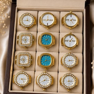Puede incluir: Una colección de doce anillos reloj de tono dorado dispuestos en una caja de madera oscura. Los anillos presentan diseños variados, incluyendo esferas redondas y cuadradas, algunas con fondos blancos o azules y otras con cristales.