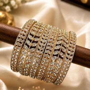 Peut inclure: Un ensemble de cinq bracelets joncs dorés présentant une rangée centrale de motifs en forme de feuilles incrustés de cristaux clairs, bordée de deux fines bandes de petites pierres scintillantes de chaque côté, formant un bijou orné.
