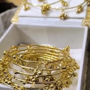 May include: Gold bangles with dangling charms, presented in a white box. The bangles are stacked, highlighting their detailed design. The charms are small, round beads. The jewellery is displayed on a white surface.