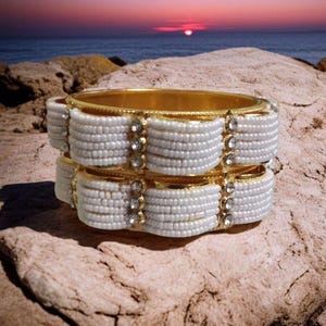 May include: Two gold bangles with white beaded accents and clear crystal embellishments. The bangles are stacked on a rock with a sunset in the background. The jewelry has a vintage or bohemian style.