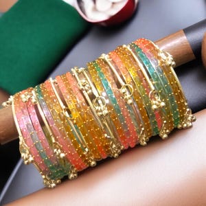 May include: A set of colorful bangles in shades of pink, orange, yellow, and green. The bangles are adorned with gold-colored accents and small dangling charms. The jewelry is displayed on a wooden dowel against a neutral background.