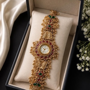 May include: A gold-toned analogue watch with a round white face featuring black numerals and hands. The watch band is highly ornate, crafted with intricate metal filigree, small dangling gold beads, and inlaid red and green oval-shaped stones.