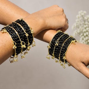 May include: Close-up of two wrists adorned with multiple black beaded bangles. Each bangle features gold-colored accents and small dangling charms. The jewelry is set against a neutral background.