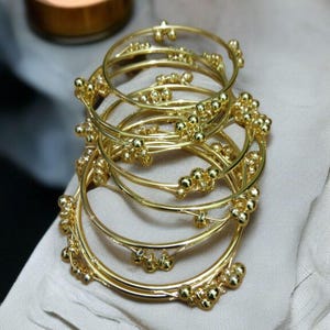 Può includere: Una pila di braccialetti rigidi in metallo color oro, ciascuno caratterizzato da una sottile fascia circolare liscia decorata con piccole perline sferiche raggruppate a intervalli regolari. I braccialetti hanno una finitura lucida e riflettente.