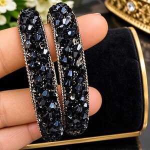 May include: Two circular bangles featuring a dense arrangement of faceted black crystals. Each bangle is framed by thin, metallic silver-toned edges with a textured, chain-like appearance, providing a contrast to the dark, reflective surface of the beads.